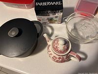 Photo showing the black Giuseppe Antonio Fratelli Dutch oven with lid, clear Fire King glass batter bowl, Churchill red and white ceramic teapot, and boxed Farberware 10-piece bowl set.