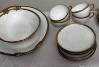 Set of white porcelain bowls with gold trim and brass handles alongside matching dessert plates and cups with gold trim handles.