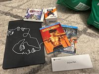 Overview photo showing all items together on carpet including Zelda 3DS game, Smartgrip in box, Birth of the Dragon and Chappie Blu-rays, Stylus Pen box, and MacBook case with line art design