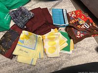 Complete view of all scarves showing a variety of colors and patterns laid out on a carpeted floor including solid burgundy, multi-colored abstract, and yellow patterned silk scarves.