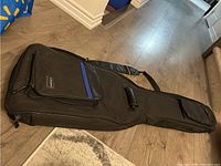 Full view of the black Voyageur soft shell guitar gig bag on the floor showing front handle, large main compartment, multiple pockets, and straps.