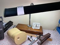 Photo shows beige vintage pencil sharpener, black retro desk lamp with wooden base, white Westclox alarm clock, metal scissors, brown stapler, and black binder on a fabric surface.