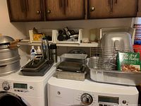 Photo of various metal baking pans stacked on a washer and dryer with some kitchen items around.