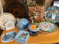 Overview of the lot showing assorted decorative plates, kettle, fabric box, candle holder, metal floral sculpture, embroidered ceramic holder, and cutting boards.