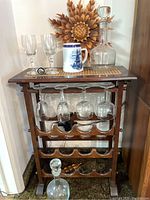 Full view of the wood and cane wine rack holding multiple wine bottles, four hanging glass goblets, a clear glass decanter with stopper, a ceramic beer stein, and a wine bottle opener. The decorative carved wooden sunburst wall piece is visible behind the rack.