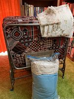 Folding cot folded upright with a mattress featuring geometric brown, black, and white patterns, floral bedding on top, and a blue and white comforter in a plastic bag leaning next to it.