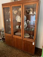 Full front view of three-door wooden cabinet with upper glass display shelves and lower solid wood storage sections, containing displayed china and silverware.