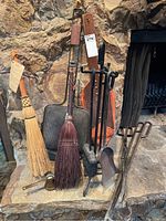 Full view of fireplace accessories arranged against a stone fireplace including two brooms, fire grate, poker, shovel, tongs, and coal scuttle.