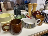 Photo shows assorted kitchen items including Tupperware bowls, ceramic mugs, glass bowls, yellow grinders, and kitchenware arranged on a white table.