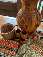 Photo showing varnished ceramic vase with carved brown patterns, four small mugs, and colorful Baltic hand-knit mittens and gloves with geometric traditional designs on a wooden table.