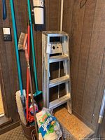Aluminum 3 step ladder next to assorted cleaning tools and paper towels on floor mat