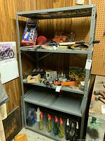 Metal shelf unit in a workshop setting against wood-paneled and pegboard walls, displaying four metal shelves holding various tools and household items (not included in sale).