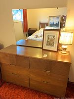 Photo showing front of mid-century wooden dresser with six drawers and large attached mirror. Table lamp and framed art piece placed on top.