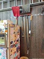 Set of three shovels hanging on wall above refrigerator, plus aluminum ladder in background