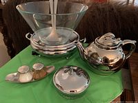 Photo showing clear glass bowl with ladle inside, polished silver teapot, silver container with lid, and salt and pepper shakers on tray