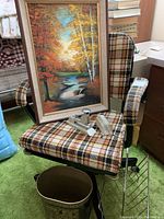 Photo showing the plaid upholstered vintage rolling work chair with original framed landscape painting resting on its seat. Also visible are the metal magazine rack next to the chair and decorative metal trash can on the green carpet.