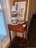 Small wooden side table with drawer and carved leg details; lamp, teacup, and framed painting on top