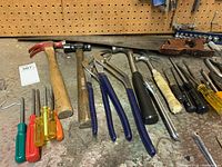 Wide view of assorted hand tools on a workbench including hammers, pliers, screwdrivers, hand saw, and files.