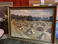 Framed painting showing haystacks in a rural field with details of houses and trees in the background, signed by the artist.