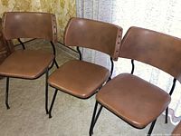 Three brown upholstered chairs with black metal frames lined up on carpet in front of yellow patterned wall and white curtain.