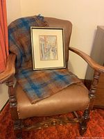 Vintage armchair in brown leather-like upholstery with carved wooden arms and legs, plaid throw blanket, and framed artwork on seat.