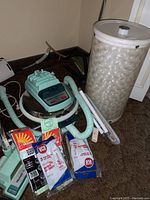 Photo showing vintage mint green Hoover vacuum cleaner with hoses, attachments, vacuum bags, and pearlized laundry hamper beside it.