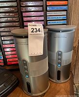 Two Centrios silver cylindrical 900MHz wireless speakers placed side by side on a wooden surface with cassette tapes in background.