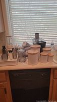 Wide view showing all items placed on kitchen counter near a window with blinds, including ceramic canisters, blender, vegetable chopper, cheese grader, ice cream maker and juicer.
