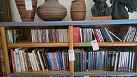 Two wooden shelves with a mixed collection of DVDs and CDs arranged side by side on both shelves with pottery and sculptures placed on top of the shelves.