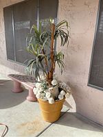 Photo of larger yellow flower pot containing a tall green artificial plant surrounded by white faux flowers. Outdoor setting on concrete patio.