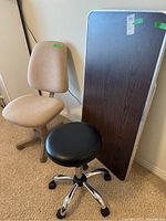 Photo showing beige office chair, black rolling stool, and brown folding table against a wall.