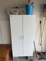 Front view of white wooden garage shelf with plastic bin and blue bucket on top, concrete blocks underneath.