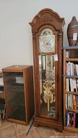Full view of the tall Ridgeway grandfather clock next to a bookshelf and wooden cabinet, showing the entire wooden case and glass front.
