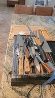 Photo of kitchen knives, forks, potato peeler, and hot plate stacked with cutting boards behind