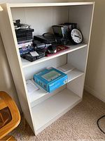 Front view of one white laminated bookcase with various objects on shelves, including clock, DVDs, boxes, and electronic accessories.