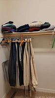 Men's dress pants hanging on wooden and plastic hangers with folded t-shirts and sweaters stacked on a shelf above.