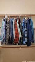 Overview photo showing 12 long sleeve button down shirts hanging on a wooden rod, displaying a variety of colors and patterns including tie-dye, plaid, and stripes.