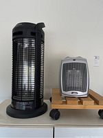 Two portable space heaters, one black Honeywell model with cylindrical shape, and one silver Lasko rectangular unit on a small wooden cart with wheels.