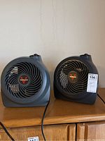Two gray Vornado fans side by side on a wooden surface with Vornado logos visible, cords attached.