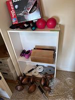 Full view of white laminate two-shelf cabinet holding pink hand weights, cardboard box, boxing gloves on top shelf, brown dumbbells and weight plates on floor.