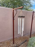 Wind chime hanging from brown painted steel base stand in outdoor gravel area against a beige wall.