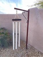 Full view of outdoor steel hanging wind chime with four silver tubular chimes and wooden top, suspended from rust-colored steel stand with decorative scrollwork, set against a stucco wall and gravel ground.