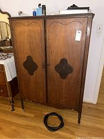 Front view of vintage wooden armoire showing two paneled doors with dark ornate panels and turned legs.