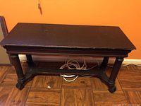 Full view of the wooden bench showing the top surface and legs against an orange wall and wood floor. Scratches visible on the bench seat.