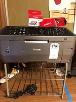 Front and top view of Canon MX870 printer on metal stand with power cord and ink cartridge boxes on top.