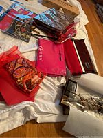 Photo showing multiple folded scarves and fabric pieces including bags and cloths in red, brown, and patterned textiles on a white cloth.