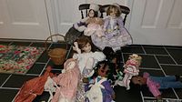 Photo showing a collection of vintage dolls seated and laid out in front of two wooden doll chairs on a tiled floor. Dolls include porcelain and plush varieties in pastel and colorful dresses.