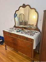 Front view of vintage wooden dresser with attached large scalloped mirror, showing decorative carved details and metal ring pulls on drawers. Several bracelets and jewelry are displayed on top covered with a white cloth.