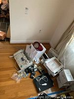 Lot of vintage electronics including Sharp fax machine, Radio Shack cell phone in box, Kindle, TI calculator and various components on hardwood floor.