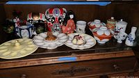 Wide view showing collection of colorful international painted masks, a traditional cloth doll, two deviled egg plates, small ceramic serving bowls with lids, two sugar bowls, and a blue floral ceramic sake set.